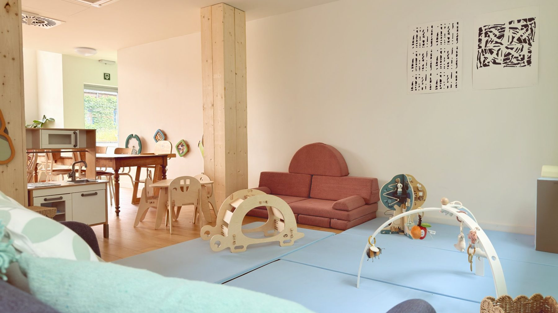 L'espace d'accueil du Village : un grand tapis de jeu bleu, un canapé en velours rose, des jeux en bois et, au fond, une cuisine d'enfant et une table près de la fenêtre